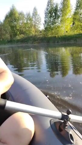 The video depicts a view from a grey inflatable boat on a calm river. A person is seated in the boat, holding an oar. The water reflects the trees on the riverbank.  A duck is visible swimming away in the distance, leaving ripples in the water.