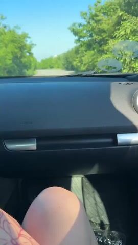The video shows a first-person perspective from inside a car driving on a sunny day in Sirakovo, Bulgaria. The camera is positioned low, capturing the car's black dashboard, silver trim, and part of a leg in the lower left corner. The leg is light-skinned, partially visible, and appears to have a subtle pattern on the clothing covering it. An air vent is visible on the right side of the dashboard. The word "AIRBAG" is clearly visible on the dashboard directly in front of the viewer.

Through the windshield, a clear blue sky is visible above a road lined with lush green trees. The road appears to be a paved, two-lane road, possibly in a rural or suburban area given the dense foliage. There are some smudges or dirt marks on the windshield. At the 0:02 mark, an oncoming dark-colored vehicle, possibly a sedan or SUV, can be seen approaching from a distance. The car continues to drive forward, and the oncoming vehicle passes by. The weather is bright and clear.
