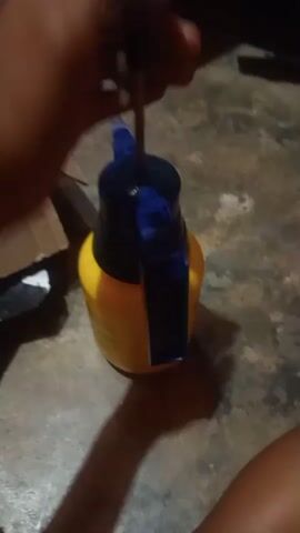 The video shows a close-up view of a hand holding a yellow spray bottle with a blue nozzle. The hand is pumping the nozzle up and down, presumably to pressurize the bottle. The background is out of focus but appears to be a floor with a textured surface. There are also some objects in the background, including a cardboard box and a black plastic bag. The lighting is dim, casting shadows on the floor. The action is slow and deliberate, suggesting the person is preparing to use the spray bottle.