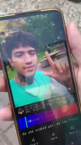 The video shows a person holding a smartphone. The phone screen displays a video of a young man with tattoos on his neck and wearing a green shirt. The man is outdoors, seemingly in a park. He initially gives a thumbs up. The camera angle of the video being played on the phone then shifts to show a group of people and motorcycles in what appears to be the same park-like setting. The footage then becomes shaky and focuses on the back of someone's head. Throughout the video, the person holding the smartphone is partially visible, and the park environment, with grass and trees, is visible in the background.