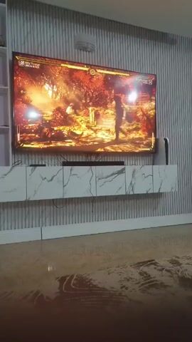 Here is a description of the video:

A video shows a television mounted on a wall, displaying a video game. The game shows two figures fighting in a forest environment. The television sits above a marble-patterned console. A Playstation 5 console is visible next to the television. The wall behind the television is decorated with vertical lines. The floor is shiny and reflects the light from the television. The camera appears to be stationary.
