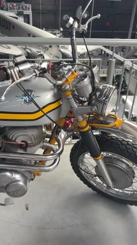 The video shows a close-up, panning view of a vintage silver and yellow motorcycle. The motorcycle is parked indoors, likely in a museum or exhibition space, with several aircraft visible in the background. The motorcycle features a silver fuel tank with yellow stripes and an MV Agusta logo, a brown seat, a yellow frame, and a chrome exhaust system. The front wheel, handlebars, and engine are also visible. The camera moves from the front of the motorcycle towards the rear, focusing on the details of its design and construction.
