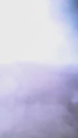 A video begins with a dark, cloudy sky. It then transitions to a solid white screen, followed by another solid white screen with a faint rectangular outline. The video then shows a ground-level view of an outdoor area. A large, light-colored rock is in the foreground, obscuring part of the view. Behind the rock, a metal grate is visible. The ground is covered in dirt and small stones, with some dry grass and twigs. Further back, a large clear plastic water bottle is situated amongst rocks. A large, dark, fluffy object, possibly an animal, is partially visible behind the rock and grate. The scene is dimly lit, suggesting it may be late in the day or overcast.