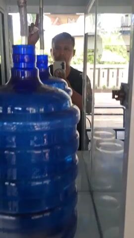 A man is reflected in a glass case, holding a phone to take a picture of himself. In front of him are two large blue plastic water bottles. A metal pipe extends from above and into the neck of the closest bottle, dispensing water. The man appears to be operating a water dispenser. The background is blurred but suggests an outdoor environment with trees and a building.