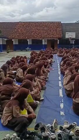 The video shows a large group of students sitting on a blue tarp in what appears to be a schoolyard. The students are wearing brown uniforms and hijabs. Most of them are looking towards the front, where a person is speaking in front of a banner. There are many shoes piled up near the edge of the tarp. In the background, there are buildings with red tile roofs and a basketball hoop. The sky is cloudy.