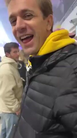 A man with blond hair, wearing a black puffer jacket over a yellow hoodie, smiles and sticks his tongue out while holding a phone to record. He is at an event with other people in the background. There is music playing and some lights. The people are wearing casual clothes and standing near each other. The interior space has a high arched ceiling.