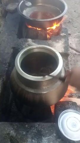 The video shows an outdoor cooking scene with two large pots over open flames. The pot in the foreground is a dark metal pot with a wide mouth, containing a dark liquid. A hand is seen stirring this liquid with a metal utensil. The pot is situated on a rough stone structure with visible burning wood and embers underneath. To the left and slightly behind, another large metal pot sits on a similar stone structure, also over flames. This pot contains a reddish-brown substance. The overall environment appears rustic and possibly rural, with natural stones and rough surfaces visible. A metal lid is placed on the ground near the foreground pot. The lighting suggests daytime, with strong shadows cast by the flames and objects. There is no visible indication of the exact location or the specific dish being prepared.