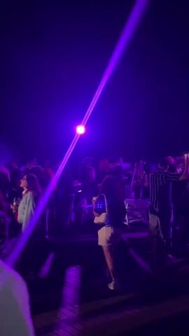 A group of people are gathered outdoors at night, illuminated by purple lights. The lighting casts strong beams and shadows across the scene. The people appear to be socializing and some are dancing. The ground is a wooden deck or platform. The overall atmosphere suggests an outdoor event or party.