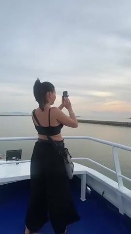 A woman stands on the deck of a boat and takes a photo of the sunrise. She is wearing a black sports bra, black pants, and a black crossbody bag. Her hair is pulled back into a ponytail. The boat has white railings and a blue deck. In the background, there is an island and a breakwater. The sky is a pale blue with pink and orange hues from the sunrise.