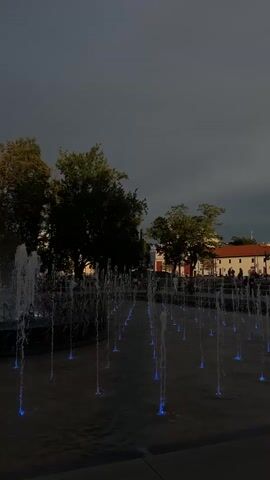 A wide shot shows a public square with a large fountain in the foreground and a building in the background. The fountain has multiple jets of water, some of which are lit with blue and purple lights. There are trees and people visible in the background. The sky is dark and cloudy, suggesting it might be evening or a storm is approaching. A bird is flying in the sky.