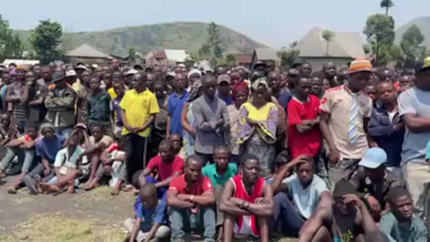 A large crowd of people, mostly men, are gathered outdoors. Some are standing, while others are sitting or kneeling on the ground. The people are of African descent. They are wearing a variety of casual clothing. In the background, there are buildings and trees. Mountains can be seen in the distance. The sky is clear and bright. Someone is speaking to the crowd in Swahili.