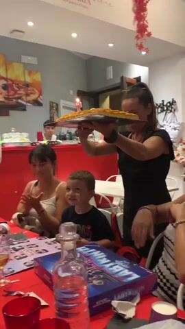 At 00:00, a woman in a black top holds a rectangular cake with a single lit candle. She is presenting it to a group of people seated at a red-covered table. A young boy in a dark t-shirt sits at the table, looking at the cake. A woman in a white top is seated next to him, clapping. A man in a white shirt stands in the background, holding a phone and looking towards the cake. The environment appears to be a restaurant, with a pizza-themed mural on the wall behind them. At 00:01, the woman with the cake continues to present it. At 00:02, the cake is still being held, and the man in the white shirt is still holding his phone. At 00:03, the woman with the cake moves it slightly, and the other people continue to look at it. At 00:04, the cake is now closer to the table, with the candle clearly visible. The people at the table are reacting to the cake.