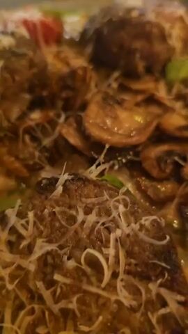 A close-up shot of a gourmet dish, possibly a type of meatball or patty, served on a dark-rimmed plate. The dish is generously topped with grated white cheese, sliced mushrooms, and small red tomato halves, resting on a creamy sauce. In the background, a wine glass filled with red wine, a bread basket, and a small gold-colored lamp are visible on a wooden table. The shot gradually zooms out to reveal a nighttime outdoor dining setting with blurred city lights and buildings in the distance.