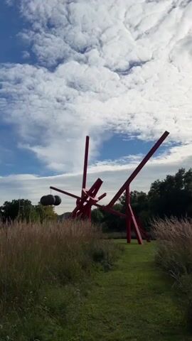 The video pans across a landscape with a large red abstract sculpture in the center. The sculpture is made of geometric shapes and appears to be constructed from metal. In the background, there are trees and a hill. The sky is blue with white, puffy clouds. Tall, dry grass and green grass are visible in the foreground and surrounding the sculpture. The camera moves slowly and steadily, offering a wide view of the scene. There are no people visible in the video. The overall impression is one of outdoor art in a natural setting.