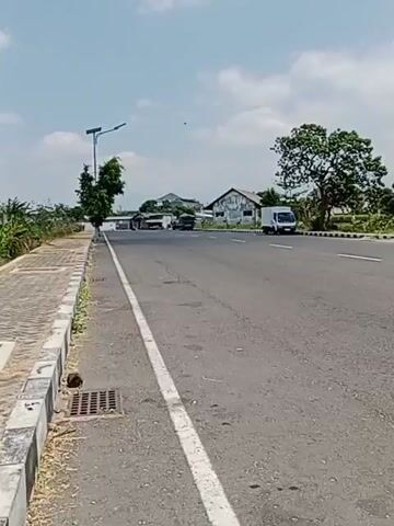 The video captures a roadside scene under a partly cloudy sky. A paved road dominates the foreground, with a white line marking the lane division. To the left, a concrete sidewalk with black and white checkered curbing runs alongside the road. A metal grate is visible on the sidewalk. Further left, a thin tree stands next to a solar-powered street lamp. In the background, several buildings with corrugated metal roofs are visible. A white van is driving on the road, moving away from the camera. A large tree with dense foliage is to the right of the road. The overall environment appears to be an open area with some vegetation.
