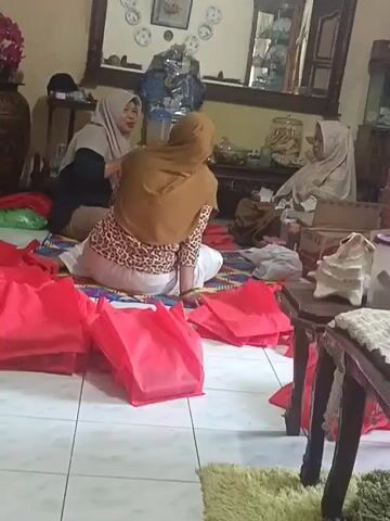 In an indoor setting with tiled flooring and a rug, three individuals are present. Two women are seated on the floor in the foreground, and another woman is seated on a chair in the background. Several red fabric bags are spread on the floor around the seated individuals. One woman in the foreground is wearing a brown hijab and a leopard-print top. The other woman in the foreground is wearing a light-colored hijab and appears to be interacting with the woman in the brown hijab. The woman in the background is wearing a light-colored hijab and is seated in a chair. The environment appears to be a living room or a common area, with furniture, shelves, and decorative items visible in the background, including a mirror above a table.