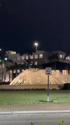 A nighttime scene shows a large illuminated fountain in the foreground, with a light-colored building visible in the background. Palm trees are planted between the fountain and the building. A "ONE WAY" street sign is visible on the right side of the frame. Cars pass by on the road in the foreground; first a white Audi and then a black pickup truck. A faint star or planet is visible in the sky.