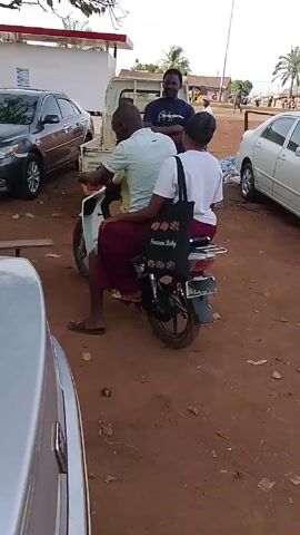The video shows a scene in an outdoor parking lot. A man is seated on a white moped with two passengers: a child sitting in front and a woman sitting behind him.  A black tote bag hangs from the back of the moped seat. Another man stands behind the moped, facing the passengers, seemingly talking to them. A fourth man walks into the frame from the right, crossing in front of the group. The parking lot is unpaved, with a reddish-brown dirt surface. Several cars are parked in the background, including a grey sedan, a white pickup truck, and a white sedan.  A low wooden bench sits in front of the moped.  A building with a red awning is visible in the background. The weather appears to be sunny.