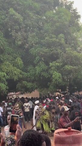 The video captures a bustling scene of a cultural event held outdoors, under the shade of a large, lush tree. A crowd of people, mostly men, are gathered around performers in traditional costumes, including masks with distinct red circular decorations, and outfits made of straw and woven materials. Many attendees are holding up their phones, recording and taking pictures of the performers. The setting suggests a village or rural area, with glimpses of simple brick structures visible in the background. The overall atmosphere is lively and celebratory, with a clear focus on documenting and participating in the cultural performance.
