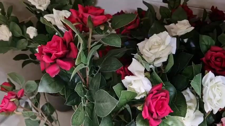 A close-up, slightly shaky video shows a dense arrangement of artificial roses and greenery. The roses are in various colors, including deep red, white, pink, and pale yellow. The leaves are a dark, realistic green. The camera pans slowly across the floral display, showing the texture and layering of the artificial flowers and foliage. The background is a neutral, light-colored wall.