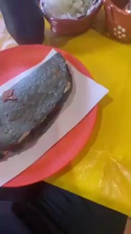 The video shows a person revealing a blue corn quesadilla on a bright orange plate, served with shredded cheese and what appears to be chorizo. A sheet of white paper towel lies between the quesadilla and the plate. The quesadilla appears well-cooked and the cheese is melted and stringy. Around the plate are other dining items, including small terracotta bowls of chopped onions and lime wedges, and possibly a green salsa. Plastic bottles of Coca-Cola and Sprite are also on the bright yellow table. The setting appears to be a casual restaurant or food stall with a brick wall visible in the background. Other people are at the table but only their hands and arms are partially visible.