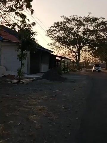 The video shows a roadside scene at sunset. On the left, there is a simple building with a covered porch. A motorcycle is parked under the porch. In the foreground, there is a pile of dark material, possibly dirt or gravel. A road runs along the right side of the scene. In the distance, there is a large tree silhouetted against the sunset sky. A white SUV is visible driving away on the road in the distance. The sky is a pale orange and yellow, indicating the sun is low on the horizon. Power lines are visible running across the top of the frame.