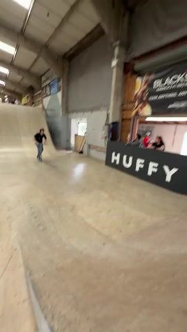 A person rides a scooter down a wooden ramp, then does a backflip in the air before landing on another ramp and riding off. The rider is wearing a black t-shirt, blue jeans, and a helmet. The ramps are in an indoor skatepark with signs and advertisements on the walls. There are other people visible in the background watching.