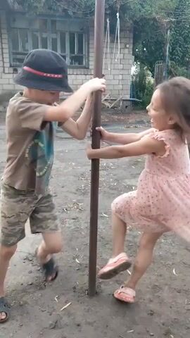 The video captures two young children, a boy and a girl, engaged in play around a metal pole in an outdoor setting. The boy, wearing a black hat with a red stripe and a tan t-shirt with camouflage shorts, is positioned on the left. He is seen holding onto the pole and appears to be spinning or pulling himself around it. His body is angled towards the girl. The girl, on the right, is wearing a light pink dress with a polka dot pattern and pink sandals. She is also holding onto the pole, mirroring the boy's actions. Both children are smiling and seem to be interacting playfully with each other as they move around the pole. The background consists of a brick building with a large window, dirt ground scattered with leaves and debris, and some vegetation. The overall atmosphere is one of active and joyful childhood play.