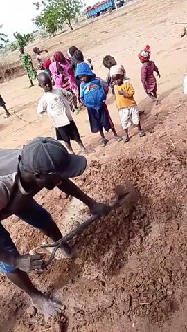 A man is digging into a pile of reddish-brown dirt with a shovel. He is wearing a gray shirt, blue jeans, a black baseball cap, and is bending over. Behind him, a group of approximately 15 children are gathered, watching. They are dressed in various colorful clothes and appear to be of different ages. The ground is a reddish, dusty color, and there are a few scattered trees in the background. A blue truck is parked farther away in the distance. There is a white bucket near the pile of dirt.