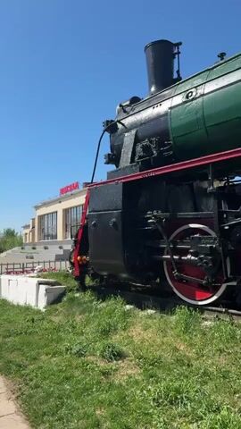 The video shows a close-up view of a green and black steam locomotive with red wheels parked on a track next to a grass area. The locomotive appears to be a historical exhibit, as there is a building with the Russian word "вокзал" (train station) in the background. The sky is clear and blue. A young child wearing a blue hat and red shirt climbs up a ladder on the side of the locomotive.