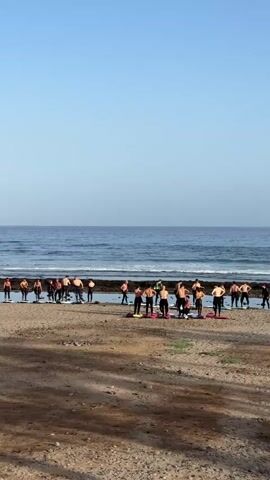 The image shows a group of people, likely an exercise or yoga class, gathered on a sandy beach in Arona, Spain. The scene is set against the backdrop of the ocean, with calm, blue water meeting a clear, bright sky. The time of day appears to be morning or late afternoon, given the soft lighting and the absence of harsh shadows.

The subjects are a diverse group of individuals, many of whom are shirtless, suggesting a warm climate and possibly a surf or beach fitness activity. They are arranged in a line or a loose formation, facing the ocean, and appear to be engaged in some form of synchronized movement or instruction. Some are standing, while others are in a kneeling or downward-facing pose. Surfboards are visible scattered among the group, hinting at the possibility of a surf lesson or a related beach sport.

The overall mood of the scene is serene and active. The natural beauty of the beach and the ocean provides a tranquil setting for the physical activity. The clear sky indicates pleasant weather conditions, perfect for an outdoor gathering. The location cues, such as the type of beach and the vegetation visible in the background, are consistent with the southern coast of Tenerife. No visible text is present in the image.