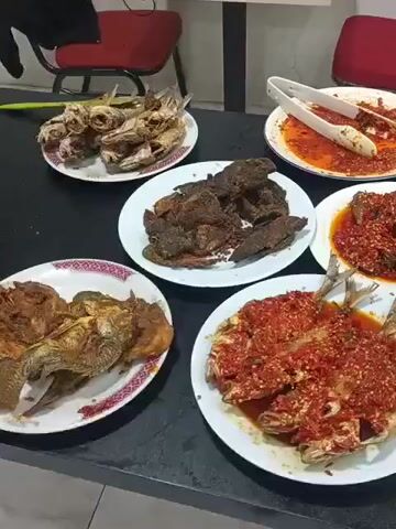 The video shows a table laden with several plates of food. The table appears to be dark, possibly a laminate or wood surface. Behind the table, two red chairs with metal legs are partially visible. The food includes several types of fish, some fried and some covered in a red sauce, as well as what appears to be fried meat and a dish of vegetables. There are also white tongs on one of the plates. In the background, a small pink basket with several light-colored eggs is visible, and a silver electronic device, possibly a fly repellent, is positioned on the table. The lighting suggests an indoor environment, possibly a restaurant or dining area. The camera is panning across the table, showcasing the spread of food.