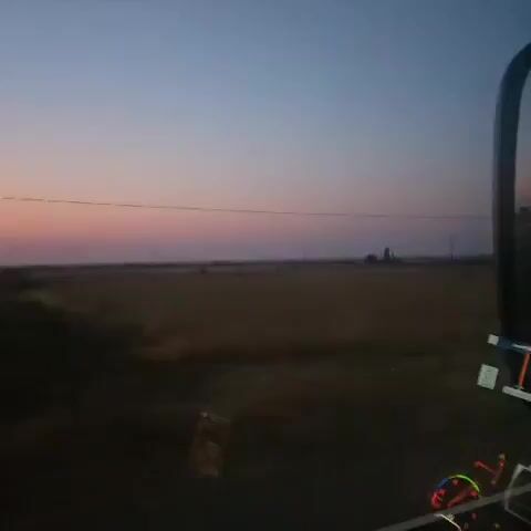 The video shows a view from inside a vehicle moving at dusk. A vast, open field stretches across the horizon, meeting a gradient sky transitioning from deep blue to pink. A thin, horizontal line, possibly a power line, is visible against the sky. A multi-story pink building with closed windows and doors appears in the middle ground. A car is parked in front of the building. Trees and foliage are visible on the right side of the frame. The interior of the vehicle is partially visible, including a dashboard with gauges.