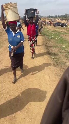 A video shows a crowd of people in a dusty, rural area. Several people are walking along a dirt path carrying buckets on their heads. These buckets appear to contain soil or dirt. The individuals are dressed in casual clothing, and there is a mix of adults and children. In the background, there is a large group of people, and a black car is parked in the distance. The sky is overcast, and the overall impression is of a busy, working environment.