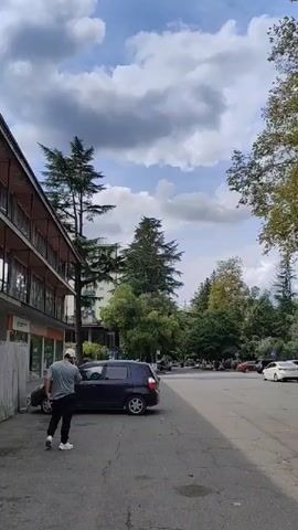 A person wearing a light-colored hat and a grey shirt walks away from the camera towards a dark-colored car parked on the left side of the frame. The car is facing away from the camera. To the right of the dark car, another dark-colored car is parked. Across the street, several cars are driving or parked. The scene is an outdoor area with trees and buildings visible in the background. The sky is partly cloudy.