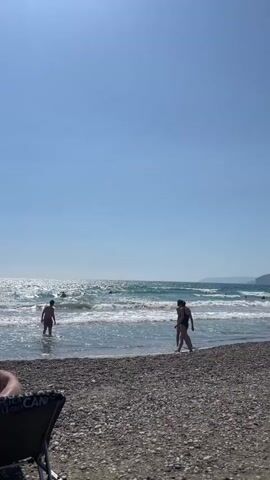 A person in a black swimsuit is standing in shallow ocean water on a pebbled beach, facing the water. Another person in a pink swimsuit is standing in the water further out, facing the same direction. Several other people are visible in the water, some appearing to swim. The sky is clear and blue, and the ocean has moderate waves. In the background, a cliff face rises from the water. In the foreground, a beach chair is partially visible.
