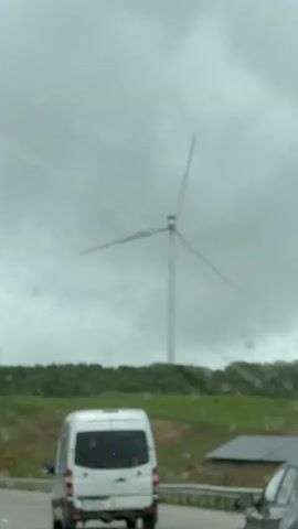 A white van is driving on a road, passing a wind turbine in the background. The sky is overcast and gray. There are some green hills and trees. The road has a barrier. There is a solar panel on the side of the road.