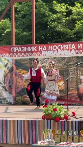 A man and a woman are performing a folk dance on a stage. The man is wearing a red vest over a white shirt, black pants with red trim, and a black hat with a red accent. The woman is wearing a white dress with intricate embroidery in red and beige, and a flower crown. They are holding hands and moving in unison. Behind them is a banner with text in a foreign language, and a backdrop depicting a rustic scene with a large pot, a spinning wheel, and a patterned rug. In the foreground, there are potted red flowers and several trophies arranged on a striped fabric. The stage is outdoors, with trees and greenery visible in the background. The lighting suggests it is daytime.