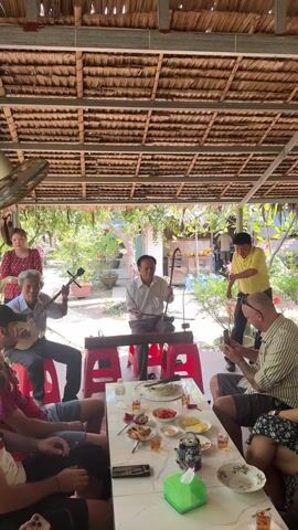 The video shows a group of people gathered in an outdoor, covered area. Four musicians are performing, with three playing instruments and one singing. Two men are seated playing stringed instruments, and a third man is seated playing a bowed string instrument.  The singer is standing, gesturing with his arms as he sings. A woman in a red dress stands behind the musicians. Several other people sit around a table, some of whom are filming the performance with their phones. The table is covered with food and drinks. The structure's roof is made of thatched materials, and a fan is attached to the ceiling. The surroundings suggest a garden or outdoor setting with greenery.