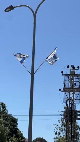 The video shows two Israeli flags attached to a lamppost, billowing in the wind against a clear blue sky.  An electrical grid tower stands to the right of the lamppost.  Trees are partially visible at the bottom of the frame.