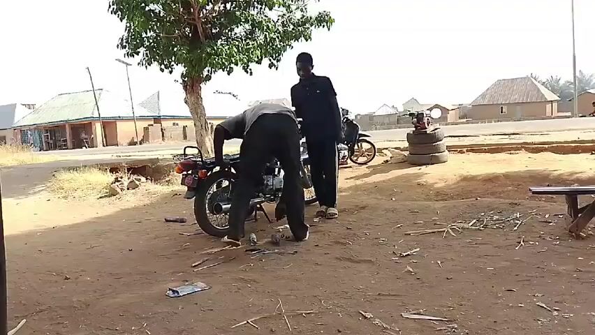 Two men are outdoors on a sunny day near a dirt area where a motorcycle is being repaired. The man closest to the motorcycle is crouched down and working on the back of the bike. There are tools and other metal objects scattered around him on the ground. The other man is standing behind him, watching. Behind them is another motorcycle parked on top of tires.  In the background, across a road, are buildings typical of a rural or semi-rural setting. A small tree provides some shade to the scene.