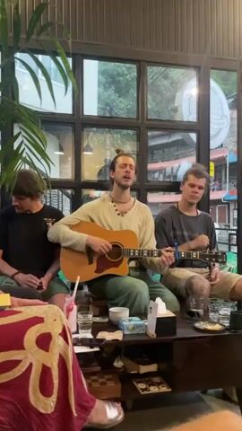The video captures an intimate musical performance taking place indoors, likely in a cafe or common area, with large, dark-framed windows offering a view of an outdoor street and lush green foliage.

Three men are present, gathered around a low wooden table. The central figure, wearing a light-colored long-sleeved shirt and green pants, has his hair tied in a bun and is adorned with necklaces. He is actively playing an acoustic guitar and singing, displaying evident passion and expressiveness. To his left, a man in a dark t-shirt sits casually, observing the performance. To the right, another man, also with his hair in a bun and wearing a striped t-shirt and light-colored shorts, sits and listens. He has a distinct tattoo on his knee and is seen at one point rubbing his face, suggesting an emotional response to the music.

On the table between them, a pink drink with a straw, several glasses of water, and a tissue box are visible. In the foreground, a vibrant red fabric with a white pattern partially covers what appears to be another seating area.

Through the large windows, natural daylight illuminates the scene, suggesting it is daytime. Outside, a round sign is partially visible, displaying text that includes "HOUSE" and "& MUSIC," hinting at the nature of the establishment. The overall mood is relaxed and engaged, characteristic of a casual musical gathering.