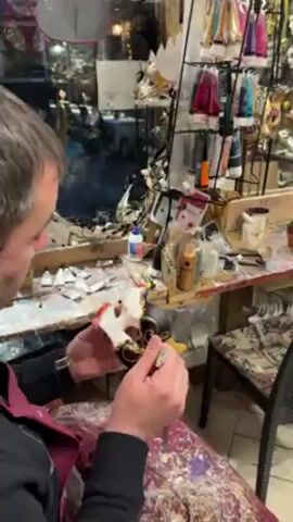 The video begins with a close-up of a man in an apron meticulously painting a mask at a cluttered workspace. He uses a small brush to add details to the black and white mask. The camera then pans to show a small seating area with black chairs and a table with a red table covering. The camera moves further into the shop, revealing two women browsing through a dense display of Venetian masks lining the walls. The shop appears to be a working studio and retail space for these handcrafted masks. The environment is packed with masks, artwork, and various decorative items.