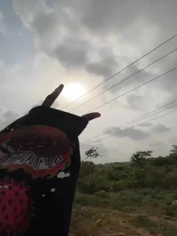 The video shows a person wearing a dark-colored sweatshirt with a graphic design on the sleeve, making a hand gesture towards the sun in a cloudy sky. The person is standing near a paved road with green vegetation and trees in the background. There are power lines visible against the sky.