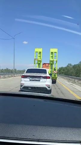 A white car is seen from behind, driving on a highway under a clear blue sky. The car has a license plate that reads B55BDV05. Ahead of the white car, a large, bright green, oversized trailer is being transported by a truck. The trailer has two tall, upright structures with multiple levels and windows. The trailer is wider than the car and takes up a significant portion of the highway lane. The environment appears to be a sunny day on a multi-lane highway, with trees and vegetation visible on the sides of the road. The video is captured from inside another vehicle, with the dashboard visible in the foreground.
