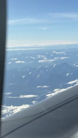 The video shows an aerial view from an airplane window. In the foreground, the wing of the airplane is visible, angled from the bottom right corner towards the center. The rest of the scene consists of a vast mountain range stretching into the distance. The mountains are snow-capped and jagged, with some clouds interspersed between them. The sky above is a clear blue with a few thin, wispy clouds. The lighting suggests it is daytime, possibly morning or afternoon. There are no people or specific actions depicted in the video, other than the implied motion of the airplane.