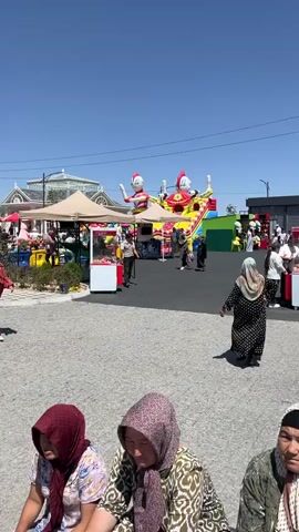 A wide shot captures an outdoor gathering with numerous people of various ages under a clear blue sky. In the foreground, several individuals are seated, facing away from the camera, some wearing headscarves. Further back, a bustling scene unfolds with people walking, interacting, and observing. A large, colorful inflatable bouncy castle with characters resembling Ultraman stands prominently in the background, along with tents and food stalls. In the mid-ground, a man in a red robe walks past a white food cart. To the right, a woman in a black and white patterned dress walks away from the camera. In the distance, a modern building with a glass dome is visible, along with a sign that reads "SHASHLIK.UZ". The ground is paved with a decorative interlocking pattern, and a metal railing separates the paved area from a grassy area with trees and flowers. The scene appears to be a public event or festival, possibly in a park or recreational area, with a mountainous landscape visible in the far background. The camera pans slightly throughout the clip, revealing more of the environment and activities.