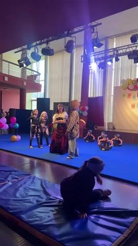 This video captures a children's performance on a stage with a bright blue floor and a backdrop featuring a stylized forest scene. Several young girls, dressed as pirates, are the central focus. Two girls on the left wear black outfits with striped leggings and sashes, while a central girl is dressed in a more elaborate pirate costume with a white blouse, a black corset, and a long, flowing red skirt. She is speaking to another individual, a taller person with curly hair wearing a plaid shirt and baggy jeans, who appears to be an adult or older teenager.

In the background, several other children, also dressed as pirates, are seated on the blue floor. Balloons in pink, yellow, and white add a festive touch to the scene. Stage lighting illuminates the performance area, suggesting it's likely an indoor event, possibly a school play or a dance recital. The mood appears energetic and engaging, with the central girl gesturing as she speaks. The image quality suggests the video was recorded indoors, possibly in a community center or school auditorium in Veliky Novgorod, Russia, given the context. The presence of a stage, lighting, and audience seating (partially visible in the foreground) confirms it's a performance setting. No specific time of day or weather conditions are discernible from the indoor scene.