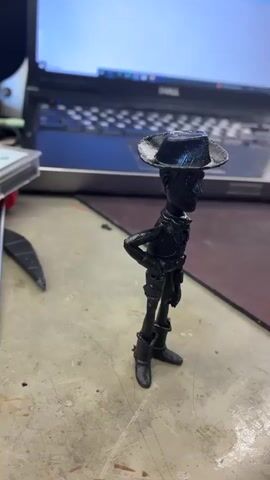 The media file is a close-up, still photograph of a black, plastic figurine resembling Woody from Toy Story. The figurine is standing upright on a workbench. Woody is wearing his signature cowboy hat, shirt, vest, pants, and boots. His left hand is on his hip, and his right arm is extended downwards. The background is blurred, showing a cluttered workbench with various tools and electronic components, including a laptop, a soldering iron, and some circuit boards. The scene appears to be indoors, likely a workshop or a hobbyist's workspace. The lighting is bright, suggesting it might be daytime, but no windows are visible. There is no discernible text in the image. The overall impression is that of a 3D-printed or painted model being worked on or displayed.