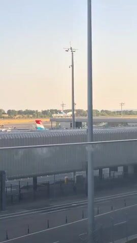 An airplane is captured mid-takeoff, its landing gear retracted as it ascends into a clear, hazy sky. The aircraft features a distinctive blue and white livery with red accents on its tail. In the background, a pink building with circular structures on its roof is visible, alongside lush green trees. The foreground is dominated by the grey, ribbed surface of an airport walkway or structure, with a tall, grey pole positioned prominently in the center of the frame. The overall setting appears to be a busy airport environment.